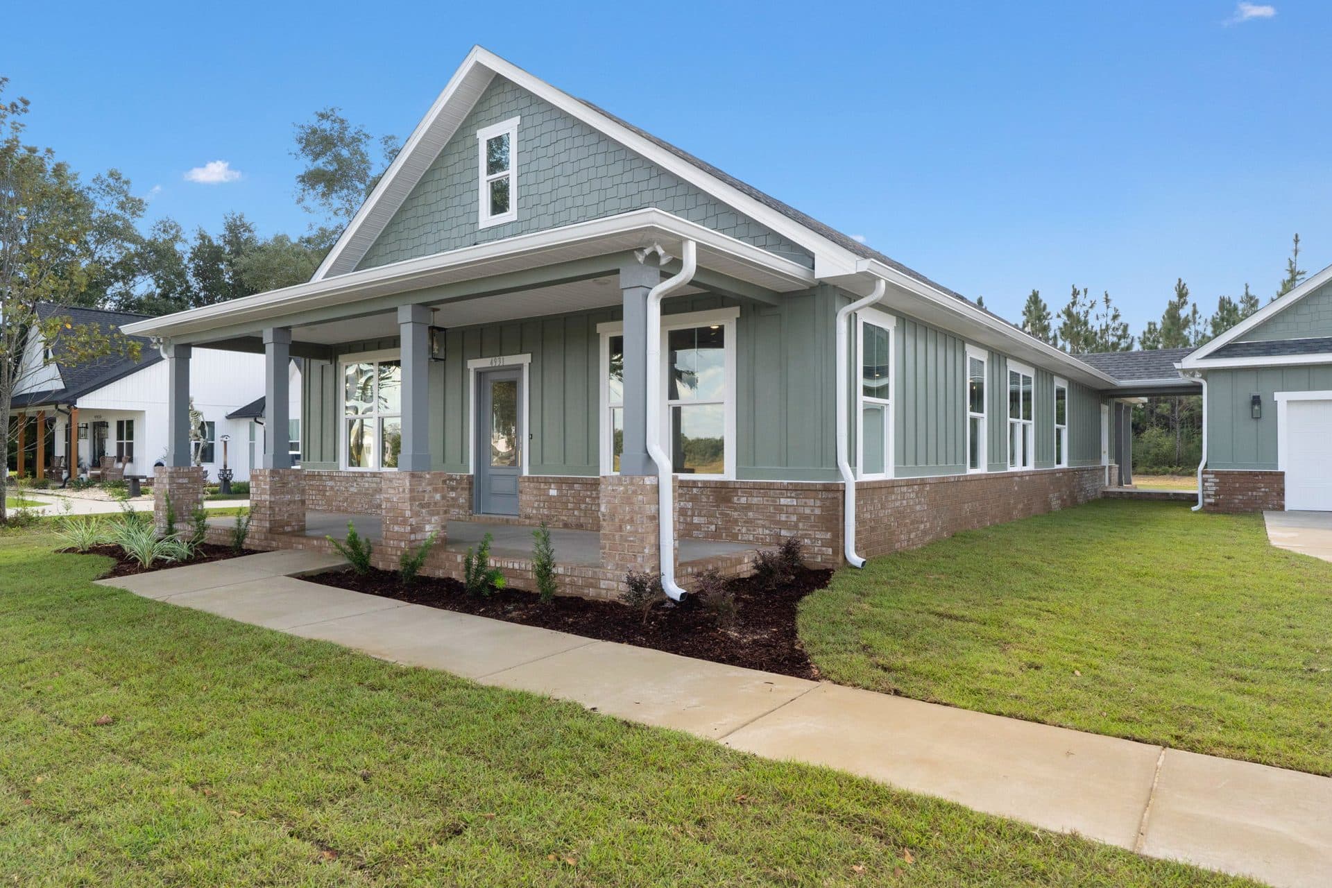 The Ashley from the front corner, sage board-and-batten over a brick water table with a deep porch and a detached 2-car garage